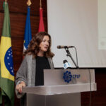 Presidnete da CIG, Sandra Ribeiro, fala num púplpito com o logótipo da CPLP. em fundo, bandeiras do Brasil, Cabo Verde e Angola