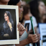 Fotografia de uma manifestação com os manifestantes desfocados a exibir um cartaz com a fotografia de Mahsa Amini