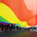 diversas pessoas pegam em conjunto numa bandeira com as cores do arco iris